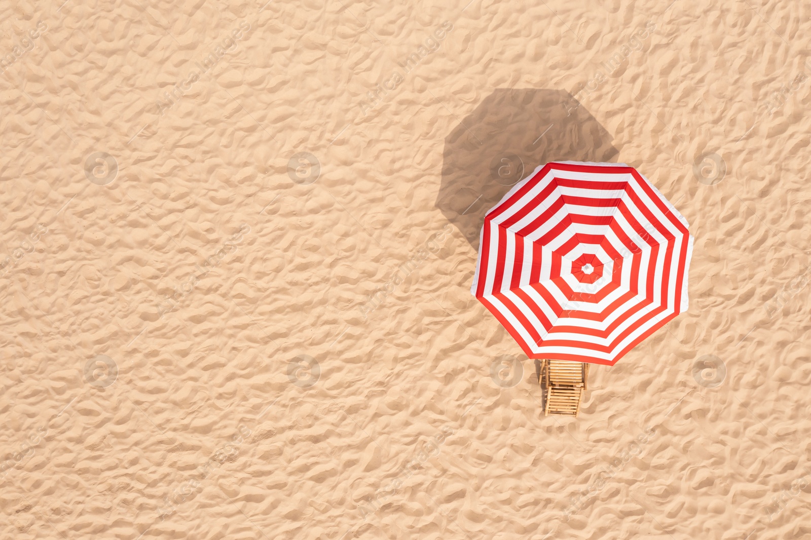 Image of Striped beach umbrella near wooden sunbed on sandy coast, aerial view. Space for text