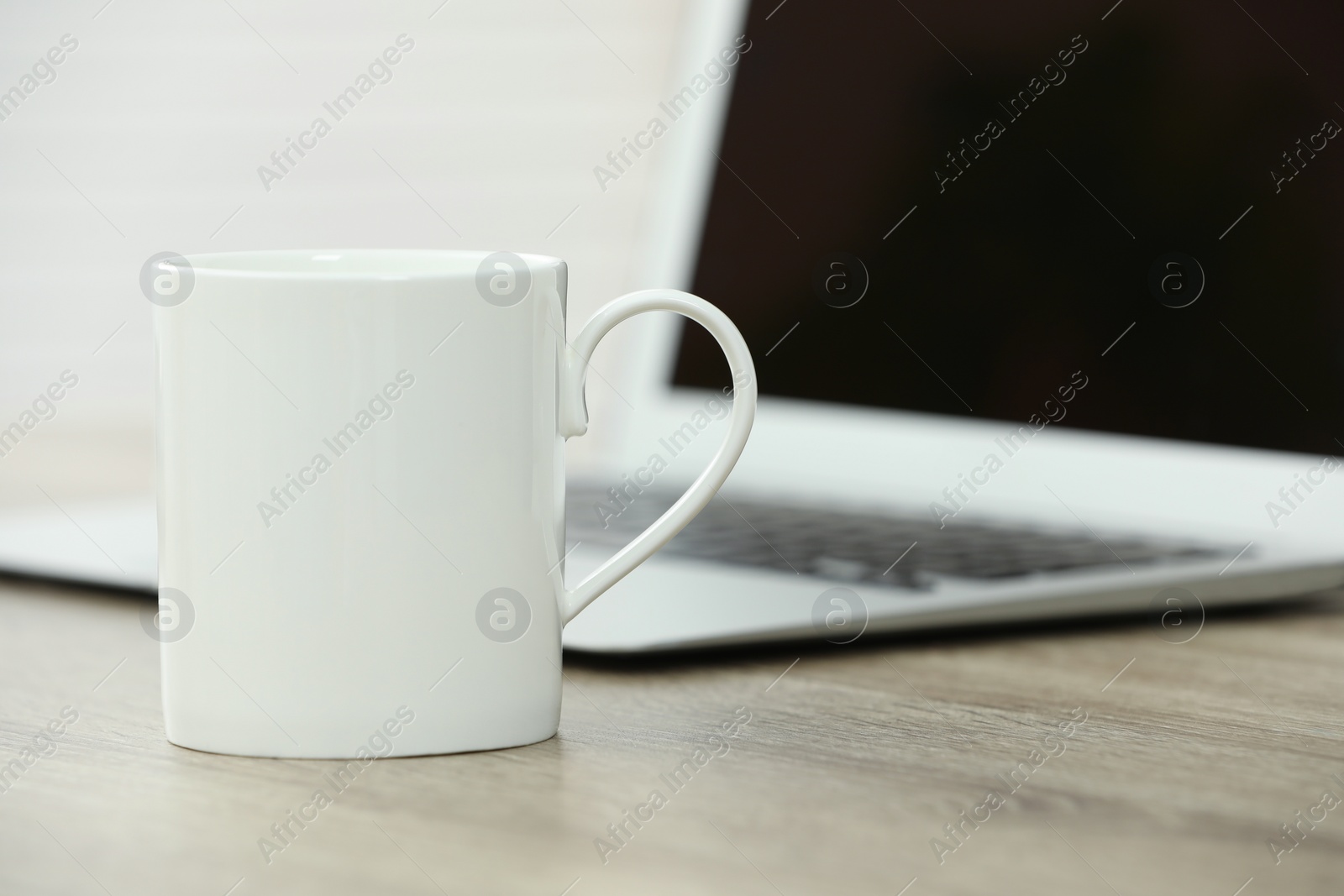 Photo of White ceramic mug and laptop on wooden table indoors. Space for text