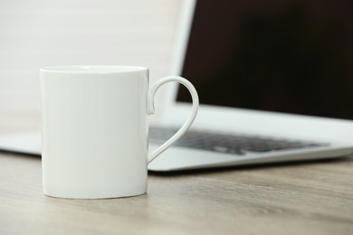 Photo of White ceramic mug and laptop on wooden table indoors. Space for text