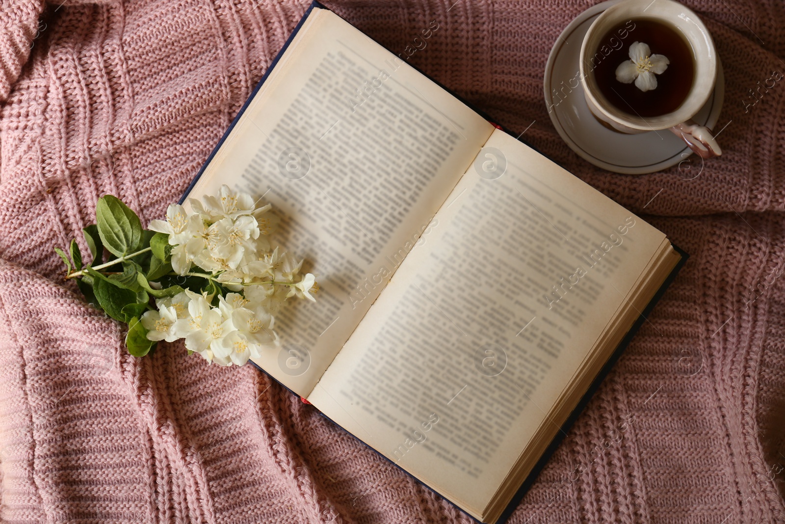 Photo of Open book, cup of aromatic tea and beautiful jasmine flowers on pink fabric, flat lay