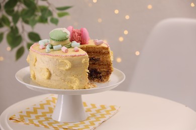 Delicious cake decorated with macarons and marshmallows on white table against blurred lights, space for text