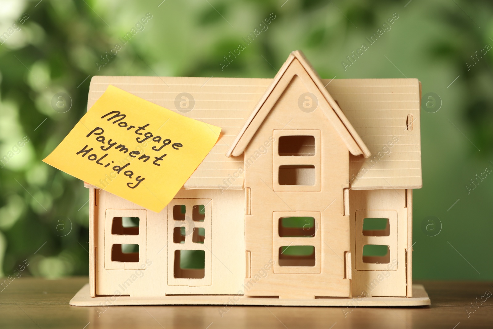 Photo of Paper note with words Mortgage Payment Holiday and house model on wooden table against blurred background