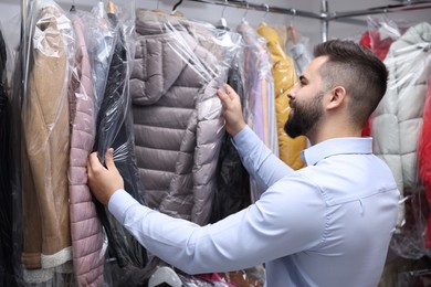 Photo of Dry-cleaning service. Happy worker choosing clothes from rack indoors