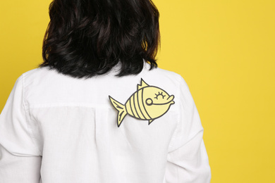 Photo of Woman with paper fish on back against yellow background, closeup. April fool's day