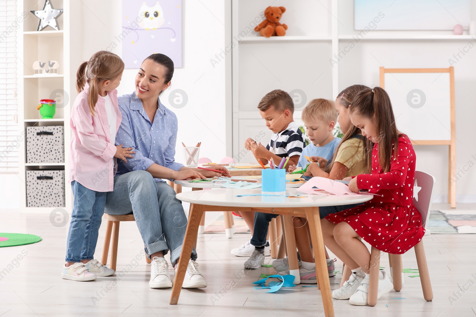 Photo of Nursery teacher and group of cute little children making toys from color paper at desks in kindergarten. Playtime activities