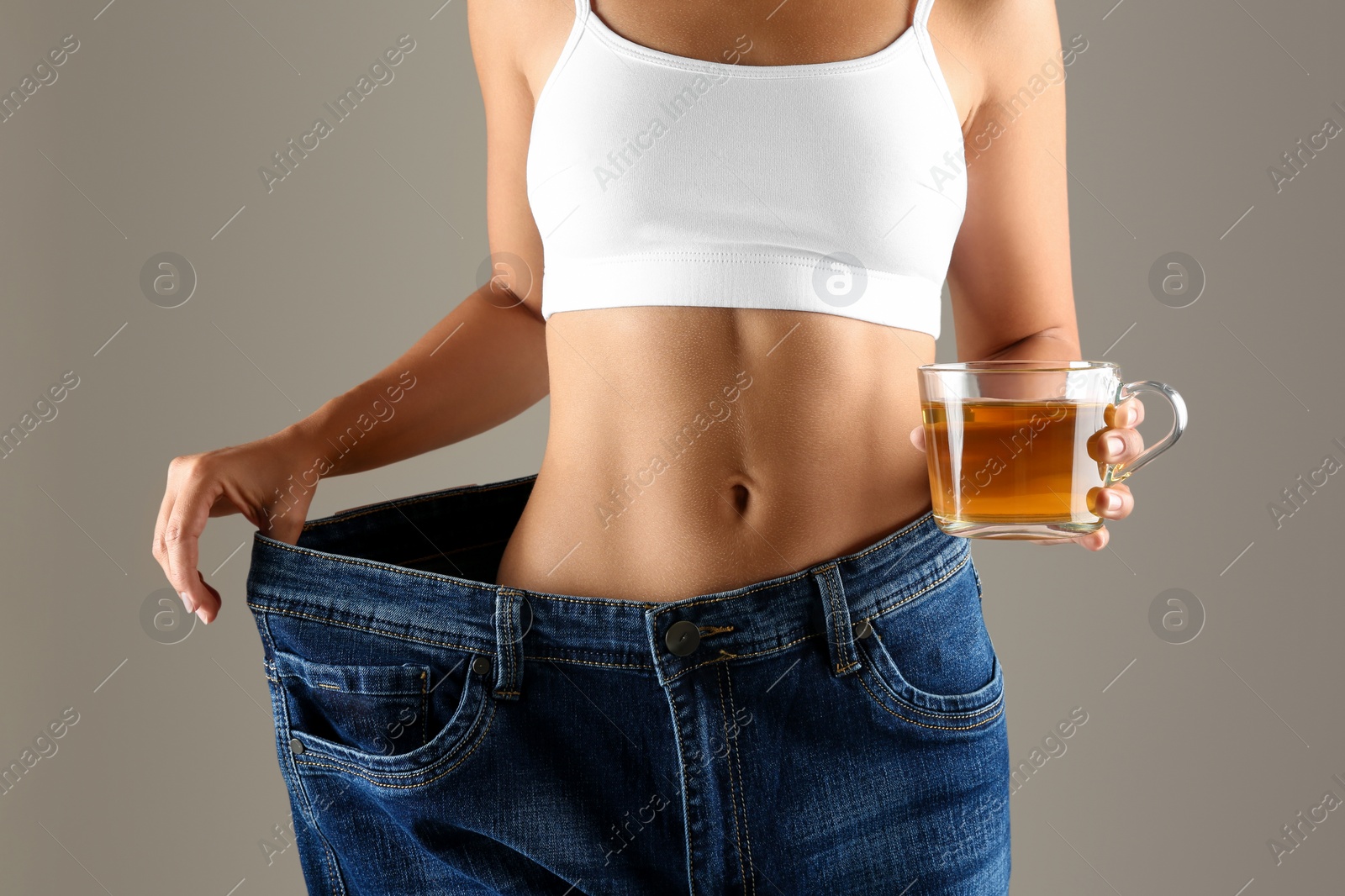 Photo of Young woman in old big jeans with cup of tea showing her diet results on beige background, closeup