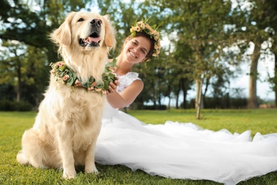 Photo of Bride and adorable Golden Retriever wearing wreath made of beautiful flowers on green grass outdoors