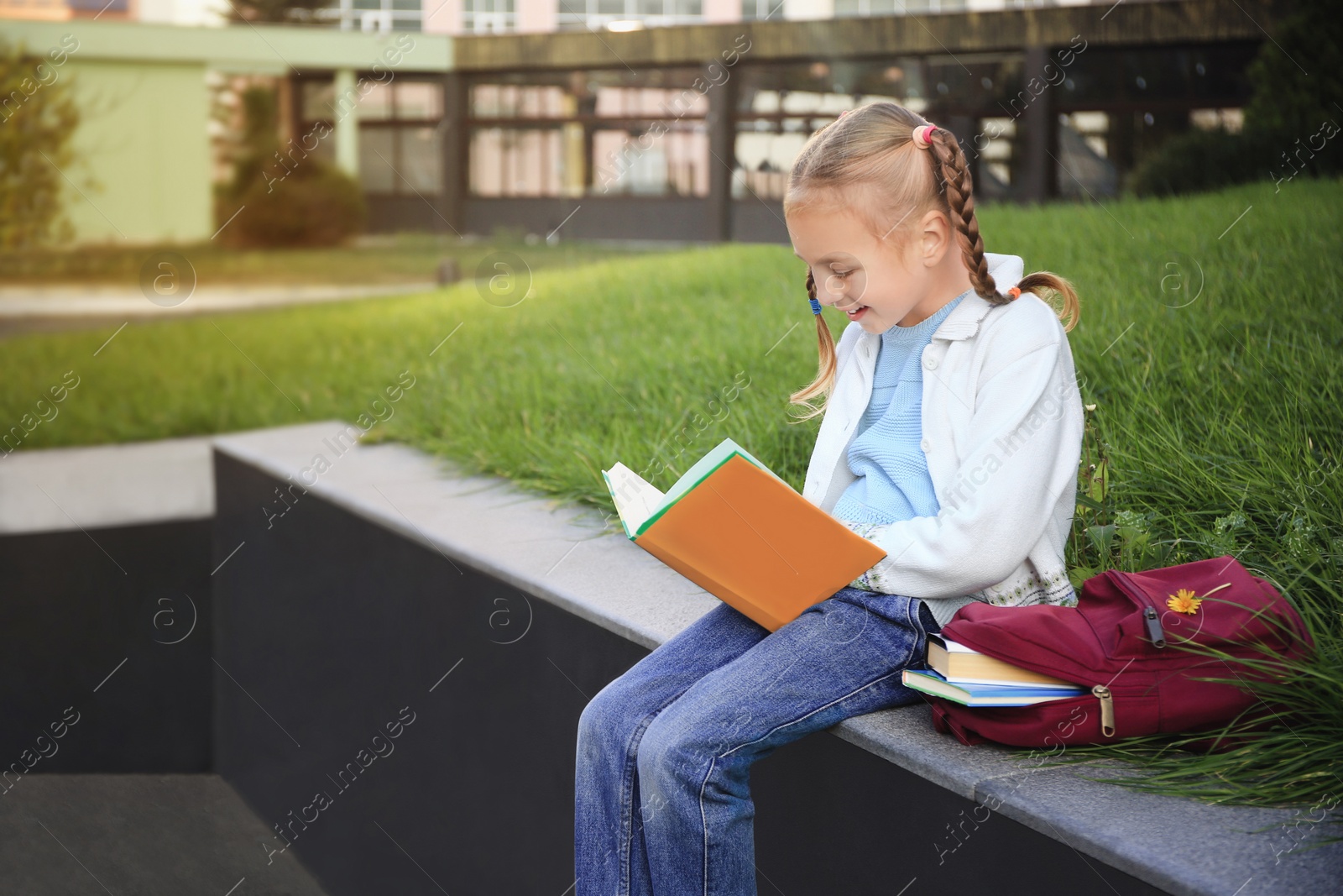 Photo of Cute little girl with backpack reading textbook on city street. Space for text