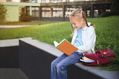 Photo of Cute little girl with backpack reading textbook on city street. Space for text