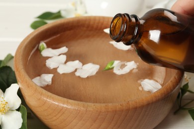 Photo of Woman pouring jasmine essential oil into bowl with petals at table, closeup