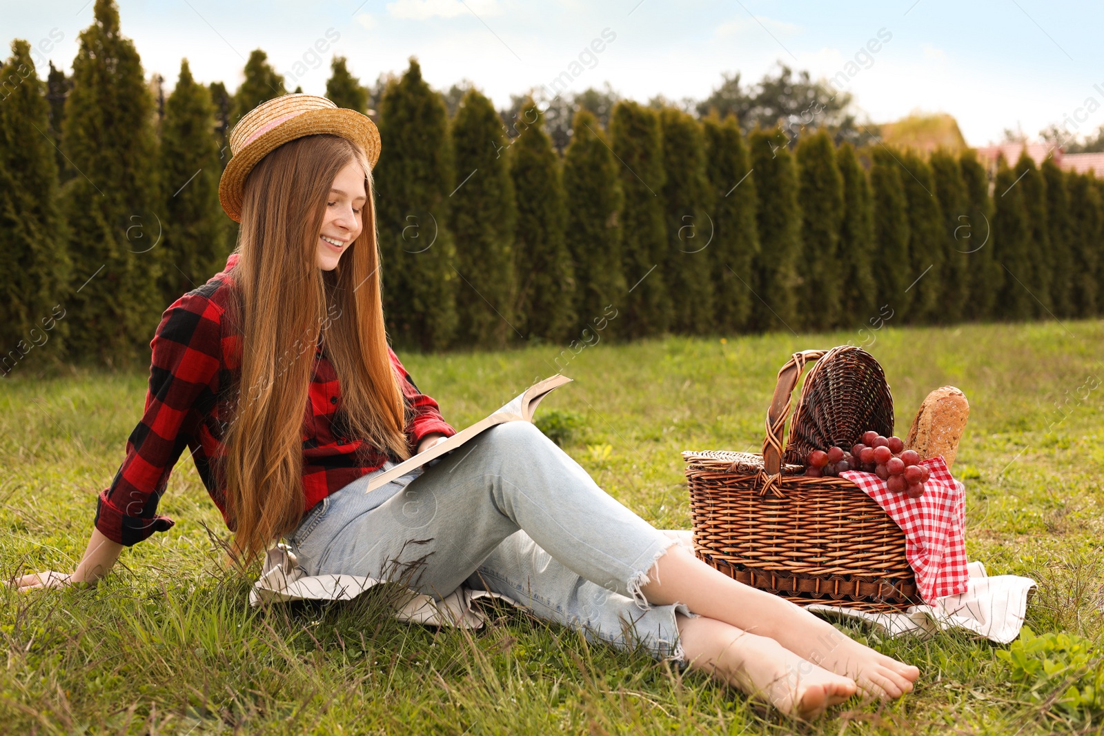 Photo of Happy girl with picnic basket reading book on green grass in park
