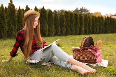 Happy girl with picnic basket reading book on green grass in park