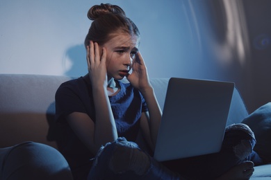 Photo of Shocked teenage girl with laptop on sofa in dark room. Danger of internet