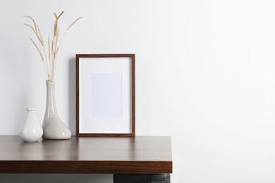 Photo of Empty photo frame and vase with dry decorative spikes on wooden table. Mockup for design