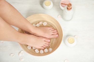 Photo of Woman soaking her feet in bowl with water and rose petals on floor, top view. Spa treatment