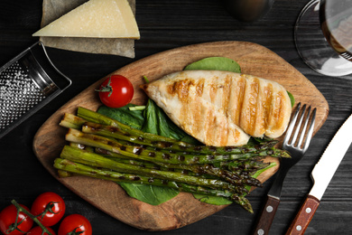 Photo of Tasty grilled chicken fillet served with asparagus on black wooden table, flat lay