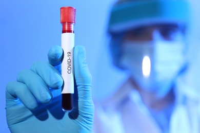Scientist in protective gloves holding test tube with blood sample and label Covid-19 against blue background, focus on hand. Space for text