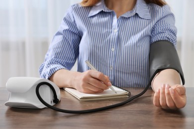 Photo of Woman measuring blood pressure and writing it down into notebook at wooden table in room, closeup
