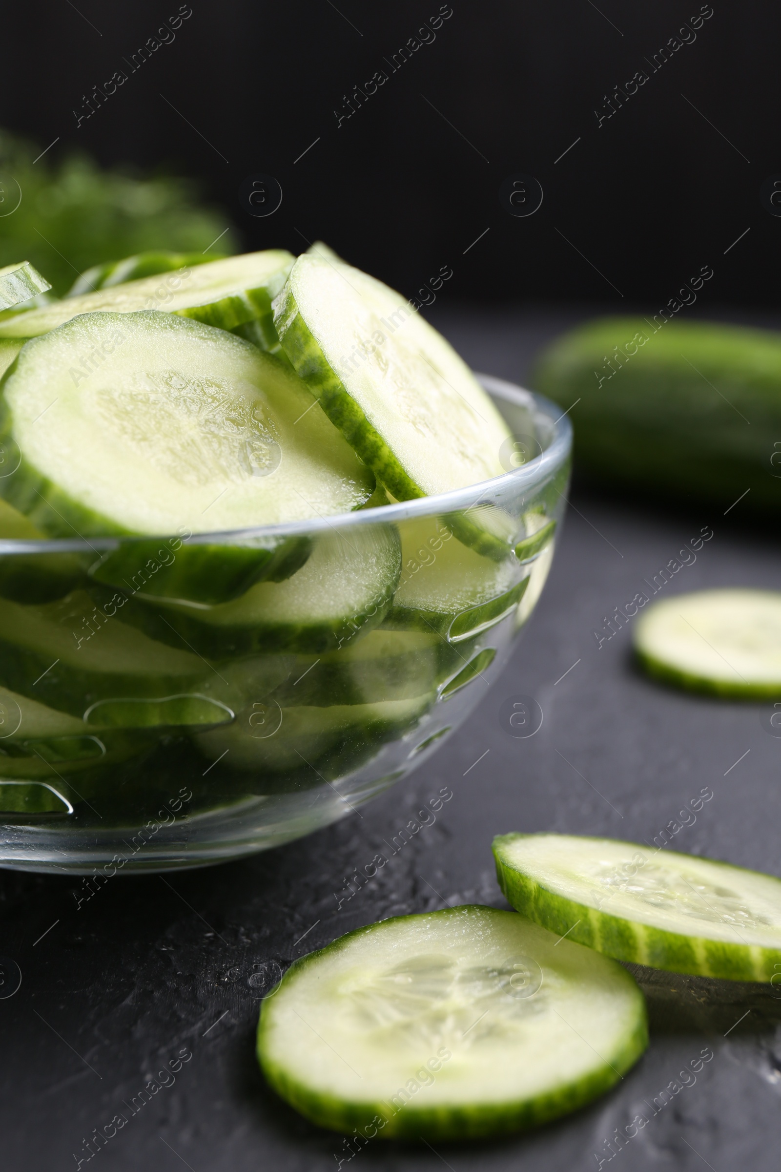 Photo of Cut cucumber in glass bowl on dark gray textured table, closeup