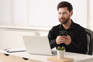 Photo of Smiling man with smartphone using laptop at table in office