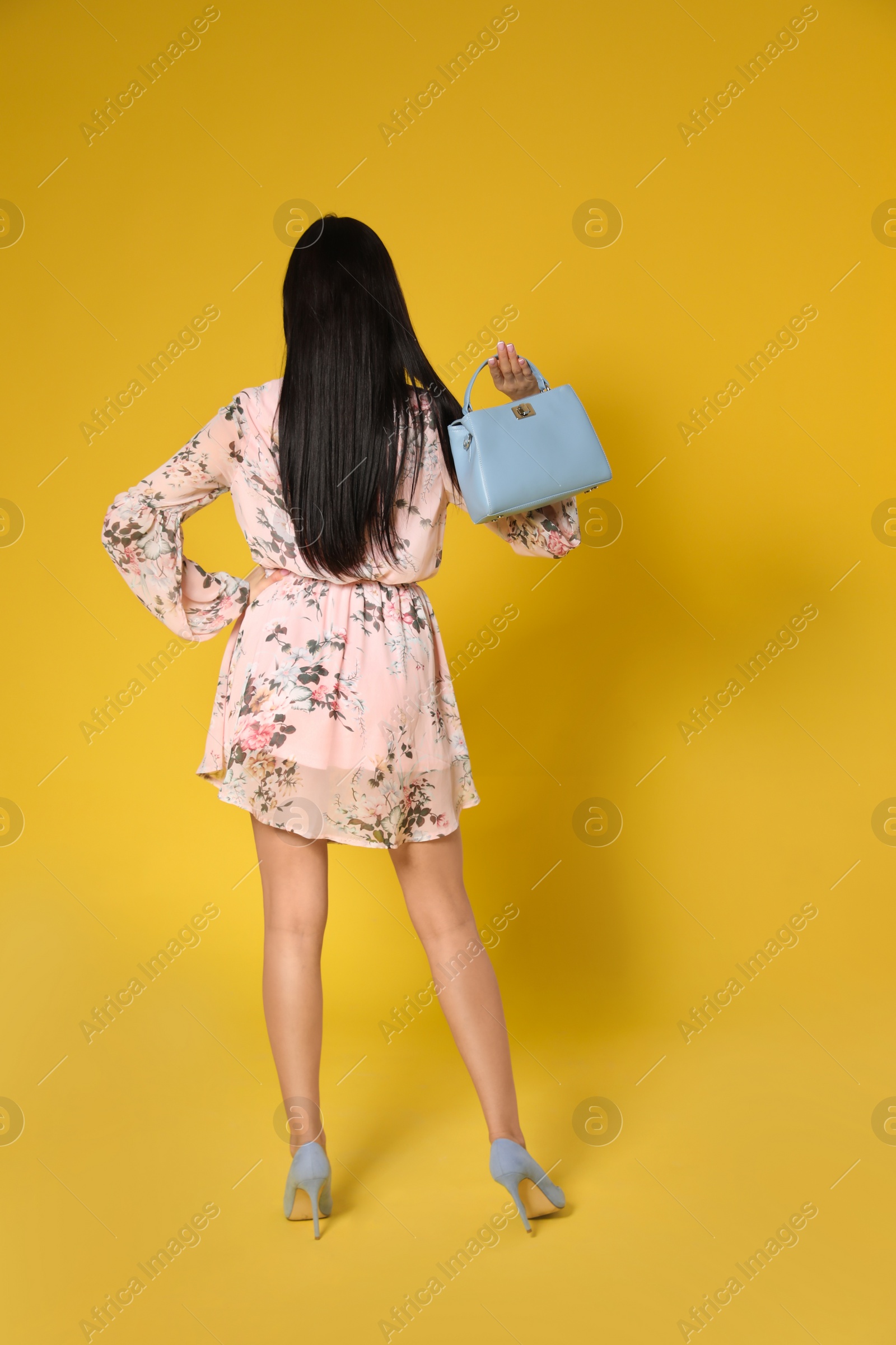 Photo of Young woman wearing floral print dress with stylish handbag on yellow background, view from back