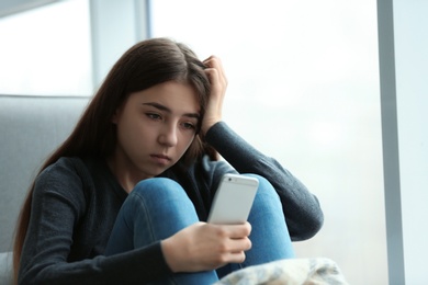 Photo of Upset teenage girl with smartphone sitting at window indoors. Space for text