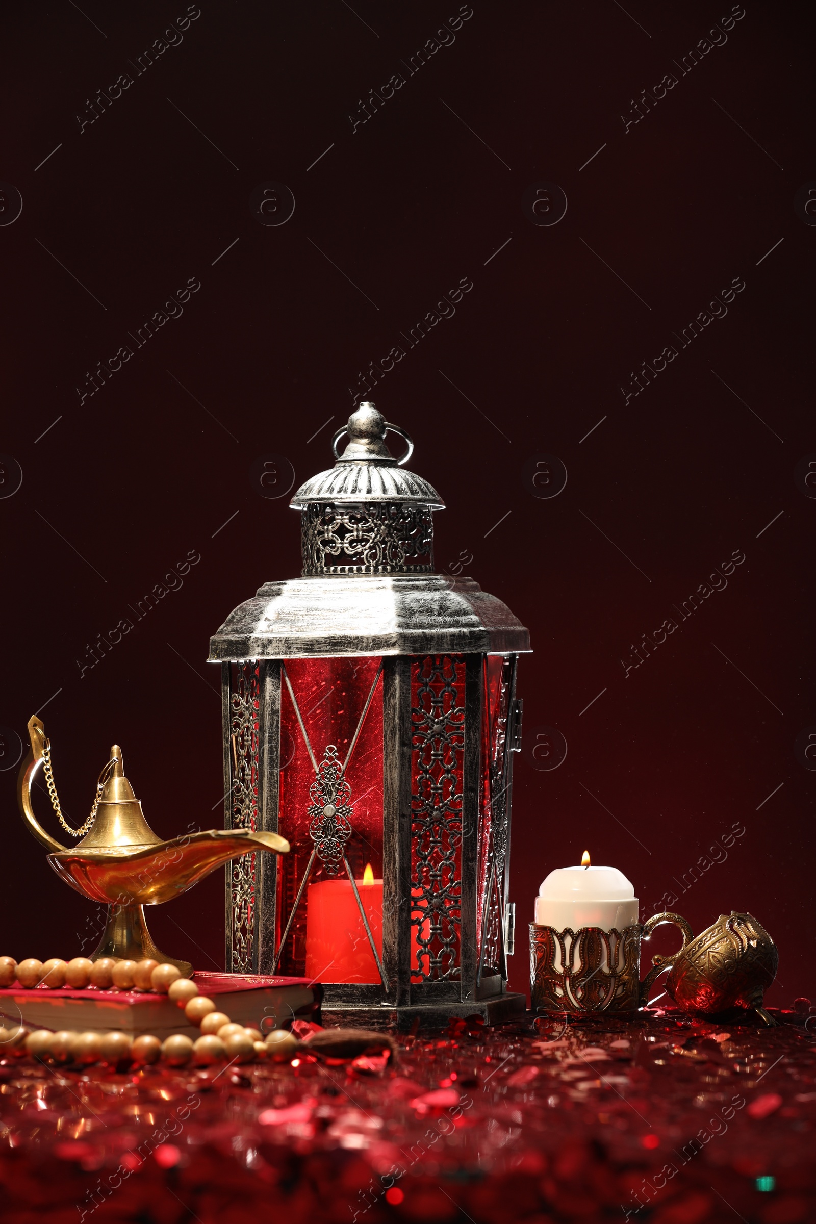 Photo of Arabic lantern, Quran, misbaha, burning candle and Aladdin magic lamp on shiny red table