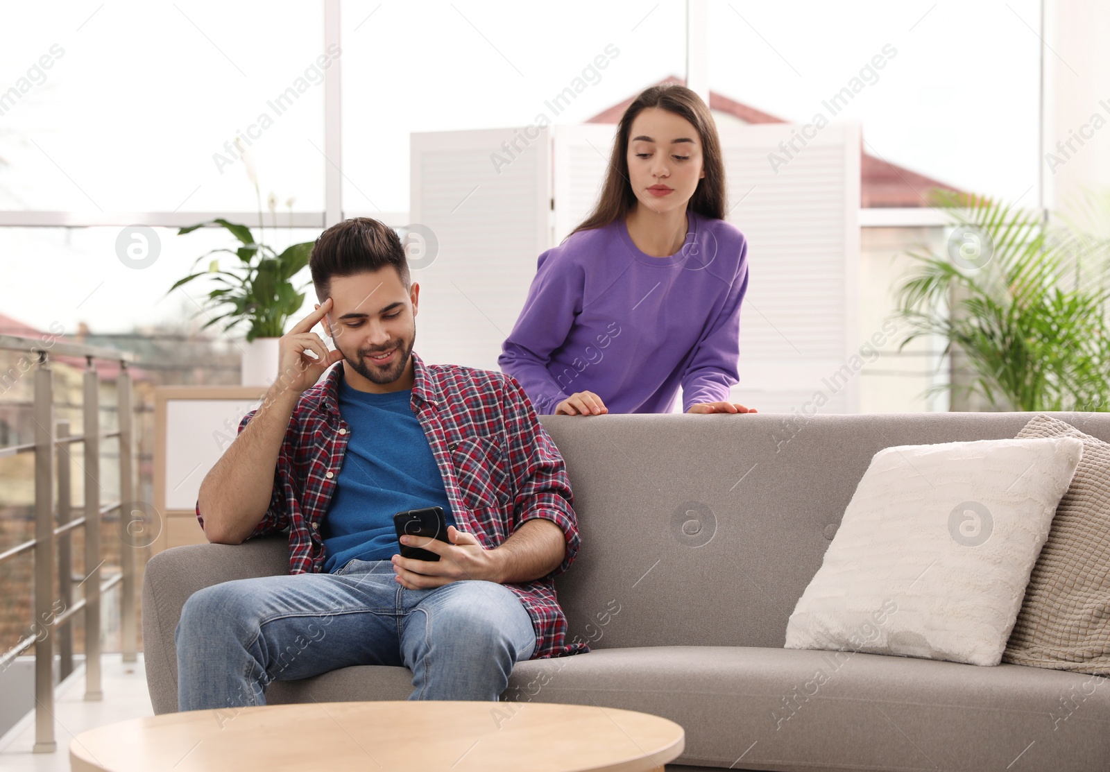 Photo of Distrustful young woman peering into boyfriend's smartphone at home