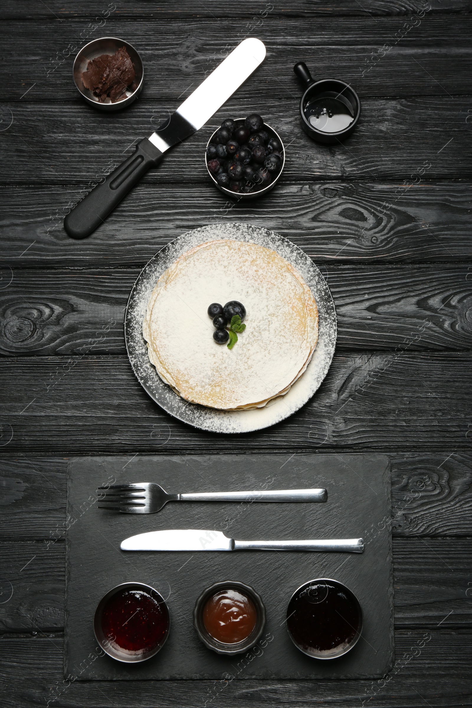 Photo of Delicious crepes, blueberries and different jams served on black wooden table, flat lay