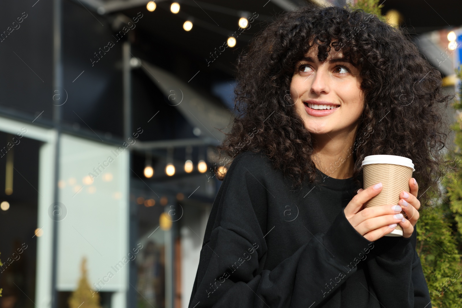 Photo of Happy young woman in stylish black sweater with cup of coffee outdoors, space for text