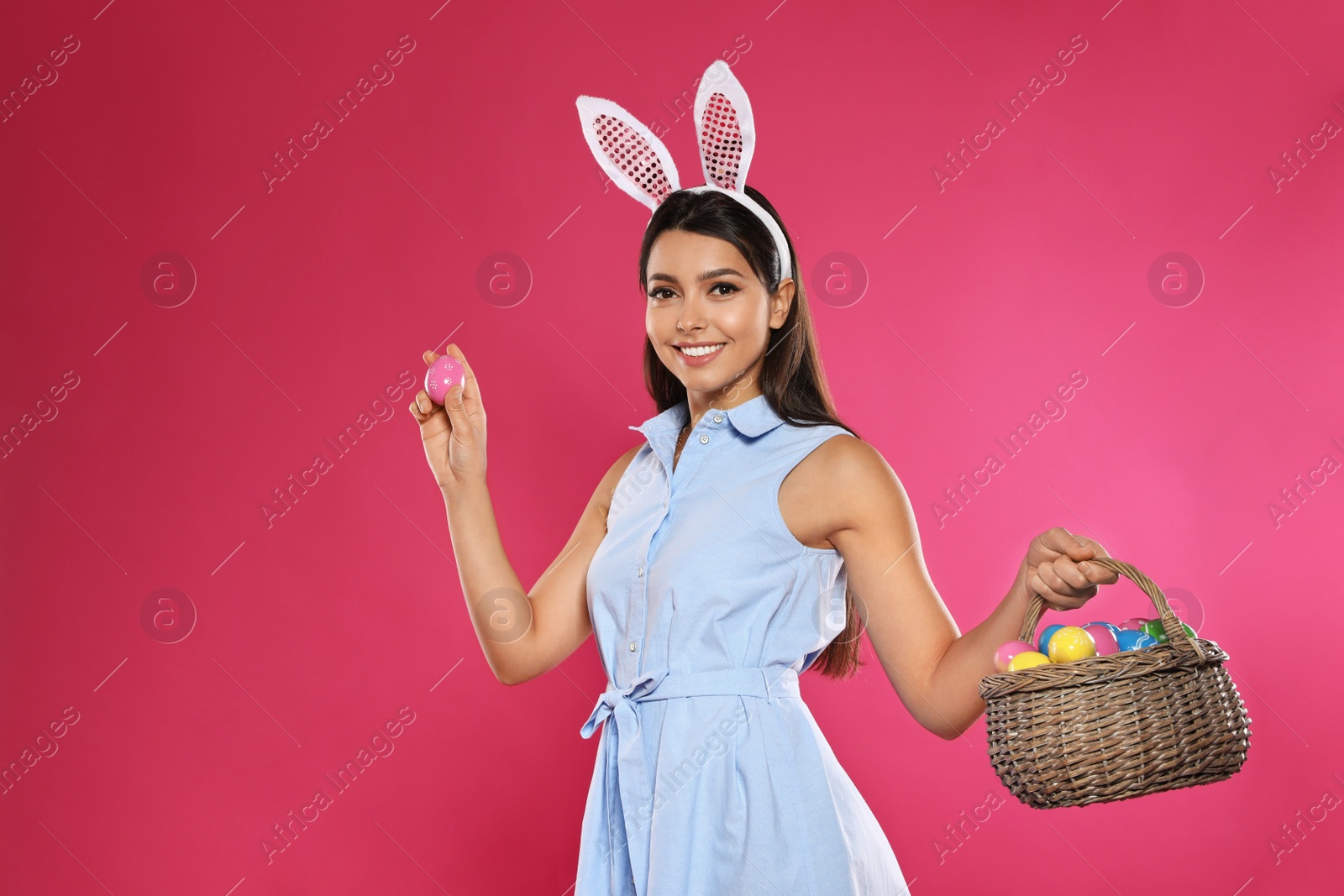 Photo of Beautiful woman in bunny ears headband holding basket with Easter eggs on color background