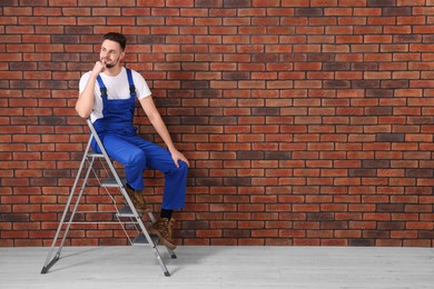 Photo of Happy worker sitting on stepladder near brick wall indoors, space for text