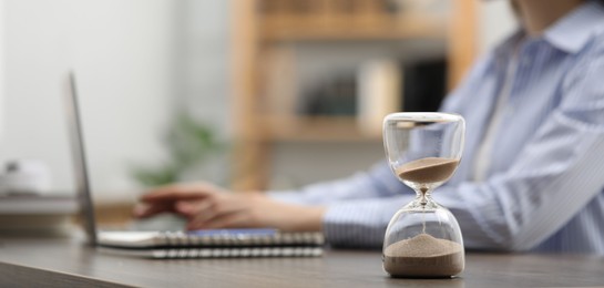 Hourglass with flowing sand on desk. Woman using laptop indoors, selective focus