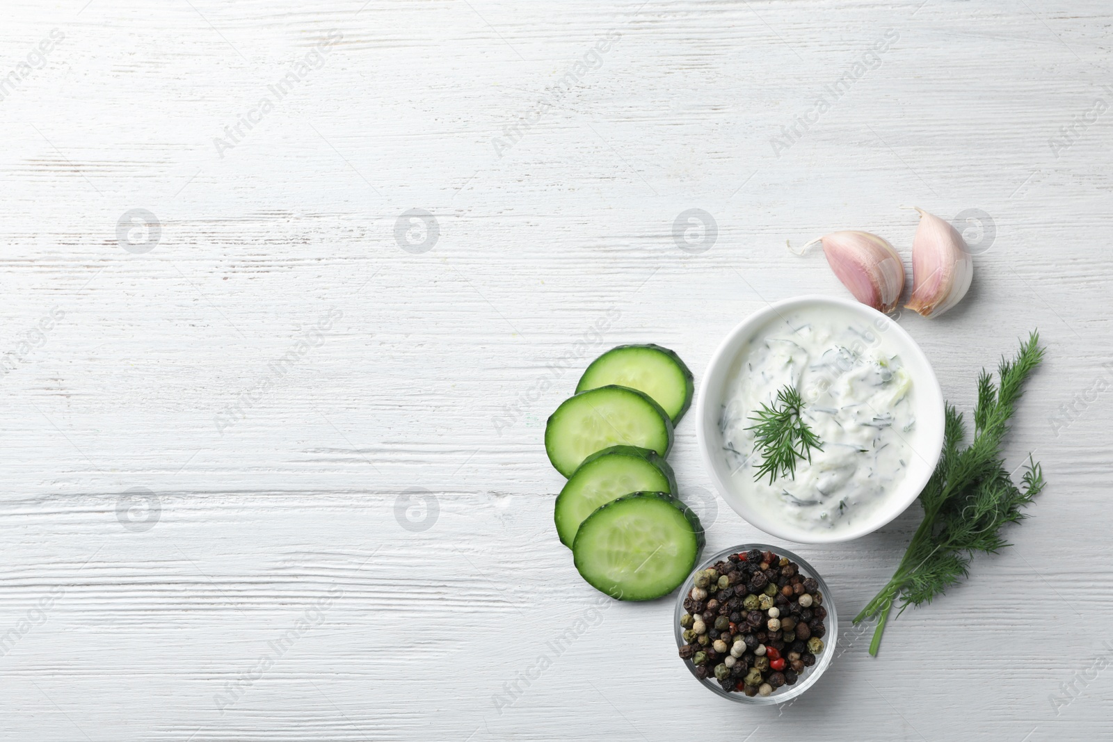 Photo of Tzatziki sauce in bowl with slices of cucumber, garlic, pepper and dill on white wooden background, top view. Space for text