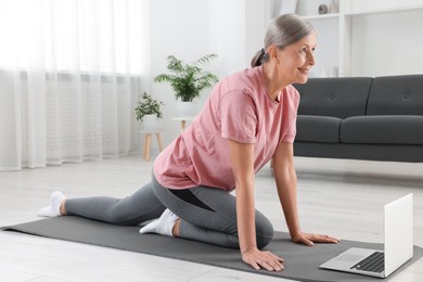 Photo of Senior woman in sportswear stretching near laptop on fitness mat at home