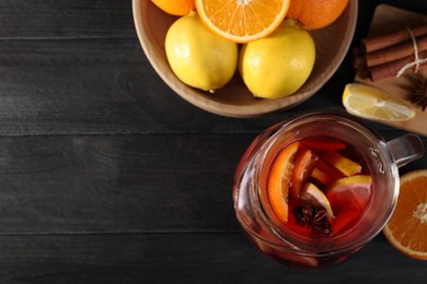 Photo of Glass jug of aromatic punch drink and ingredients on black wooden table, flat lay. Space for text
