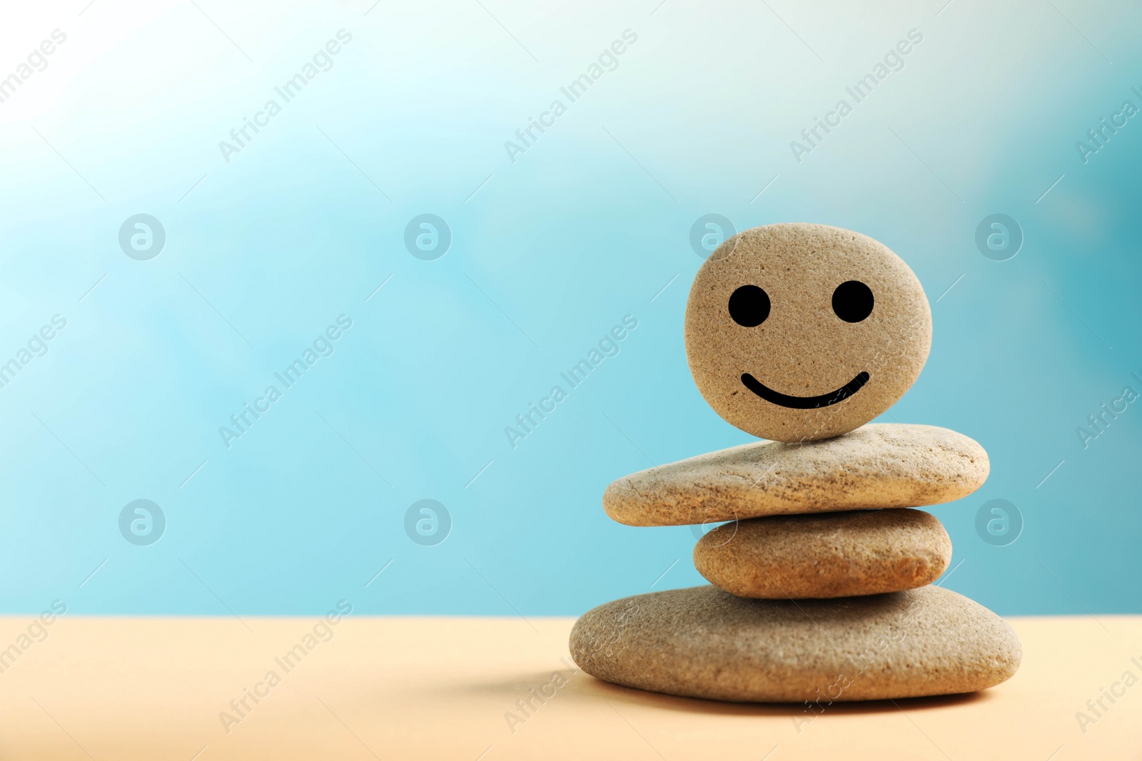 Photo of Stack of stones with drawn happy face on beige table against light blue background, space for text. Zen concept