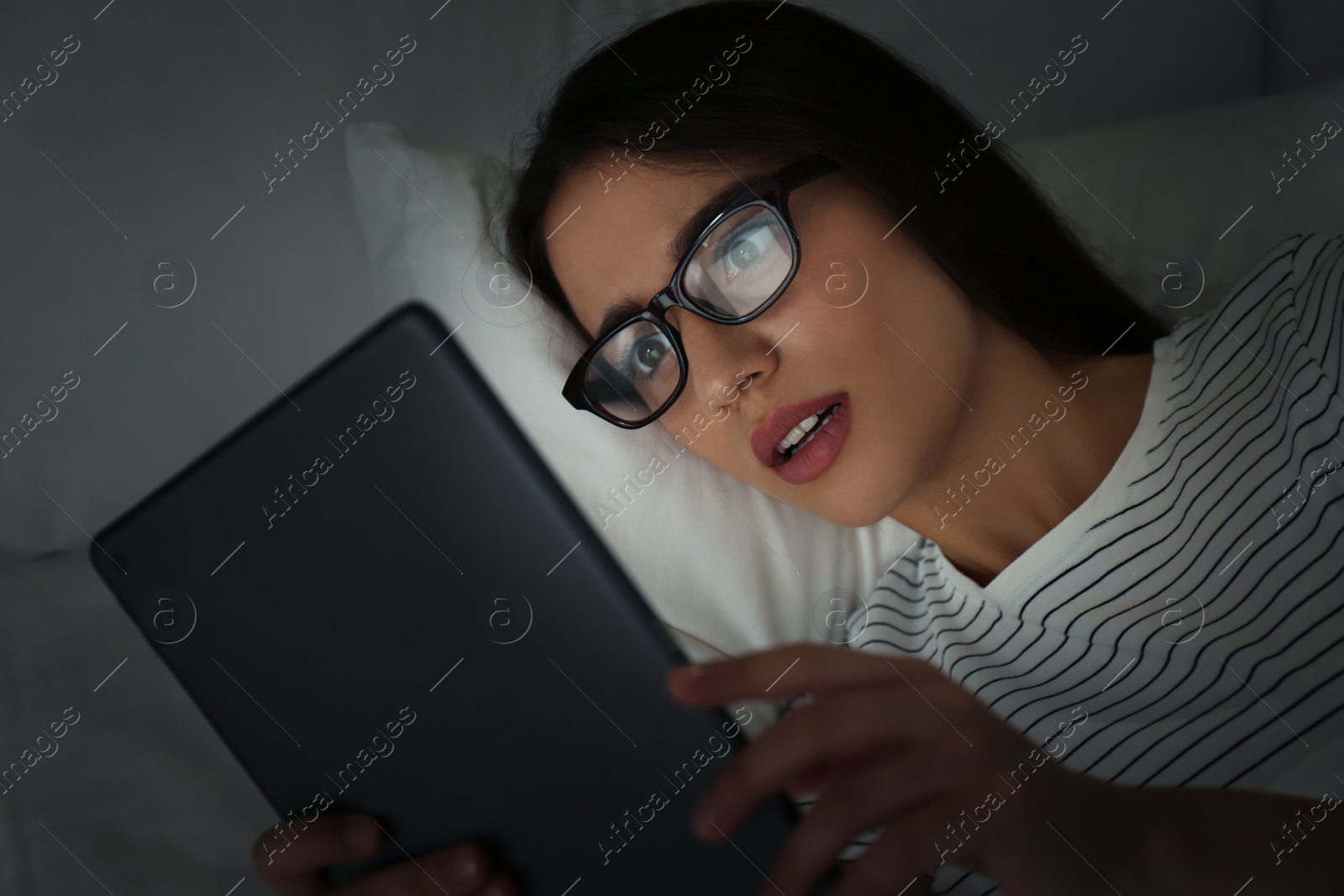 Photo of Young woman in glasses using tablet in dark bedroom