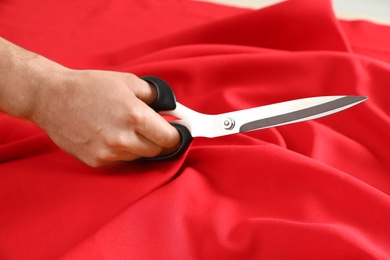 Professional tailor cutting red fabric with scissors in workshop, closeup