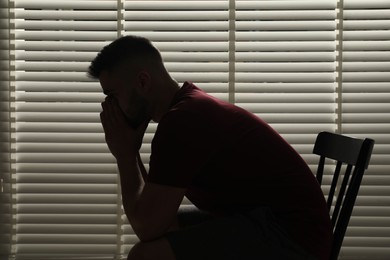 Photo of Sad man sitting near closed blinds indoors