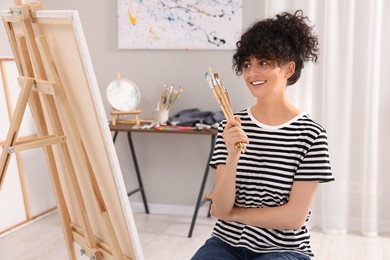 Young woman holding brushes near easel with canvas in studio