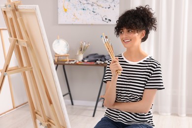 Photo of Young woman holding brushes near easel with canvas in studio