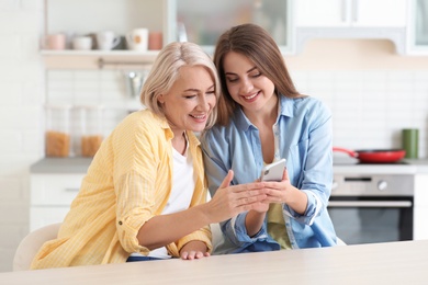 Portrait of young woman and her mature mother with mobile phone at table indoors