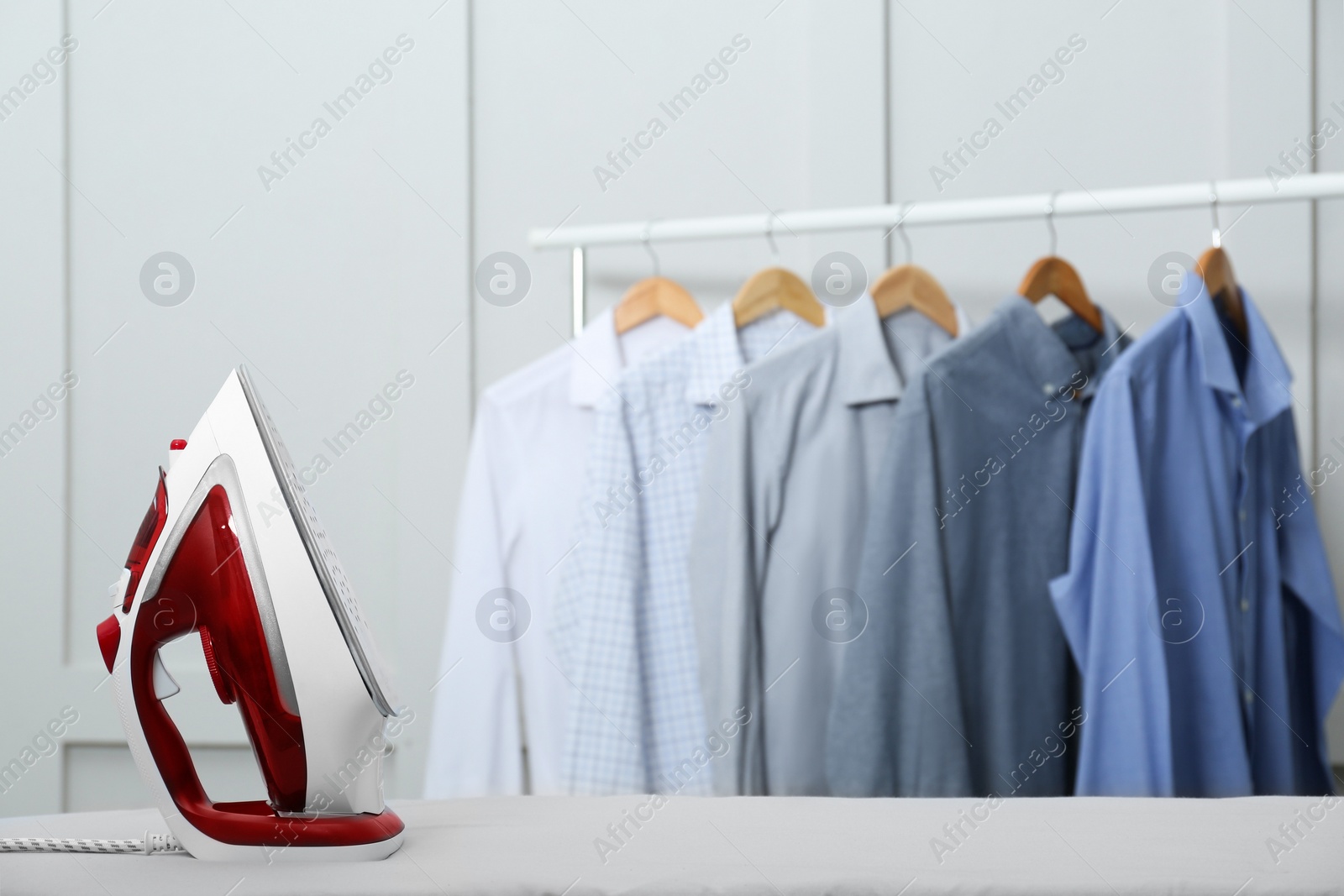 Photo of Modern iron on light table near rack with clean shirts indoors. Space for text