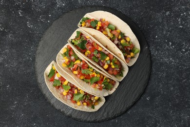 Tasty tacos with vegetables on black textured table, top view