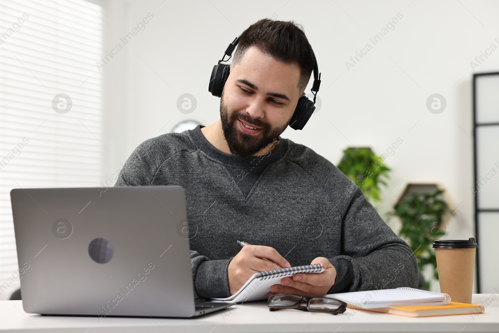 Photo of E-learning. Young man taking notes during online lesson at white table indoors