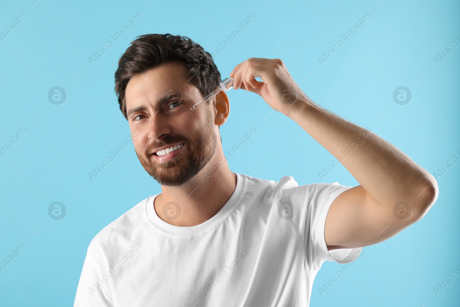 Photo of Smiling man applying cosmetic serum onto his face on light blue background