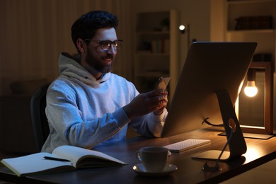 Photo of Home workplace. Happy man using smartphone while working with computer at wooden desk in room at night