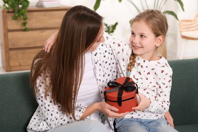 Photo of Cute little girl presenting her mother with gift on sofa at home