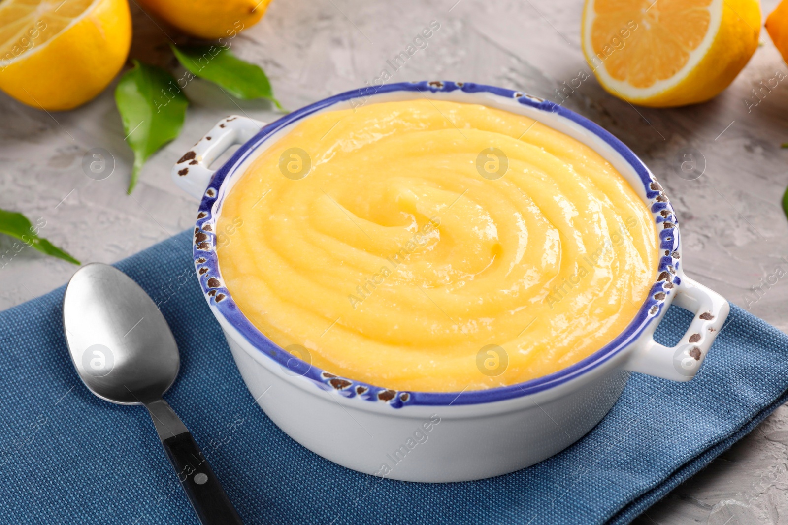 Photo of Delicious lemon curd in bowl, fresh citrus fruit, green leaves and spoon on grey table, closeup